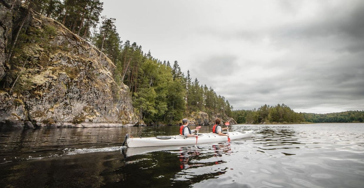 Zwei Personen paddeln in einem weißen Kajak auf einem ruhigen See neben einem felsigen, baumbestandenen Ufer unter einem bewölkten Himmel.