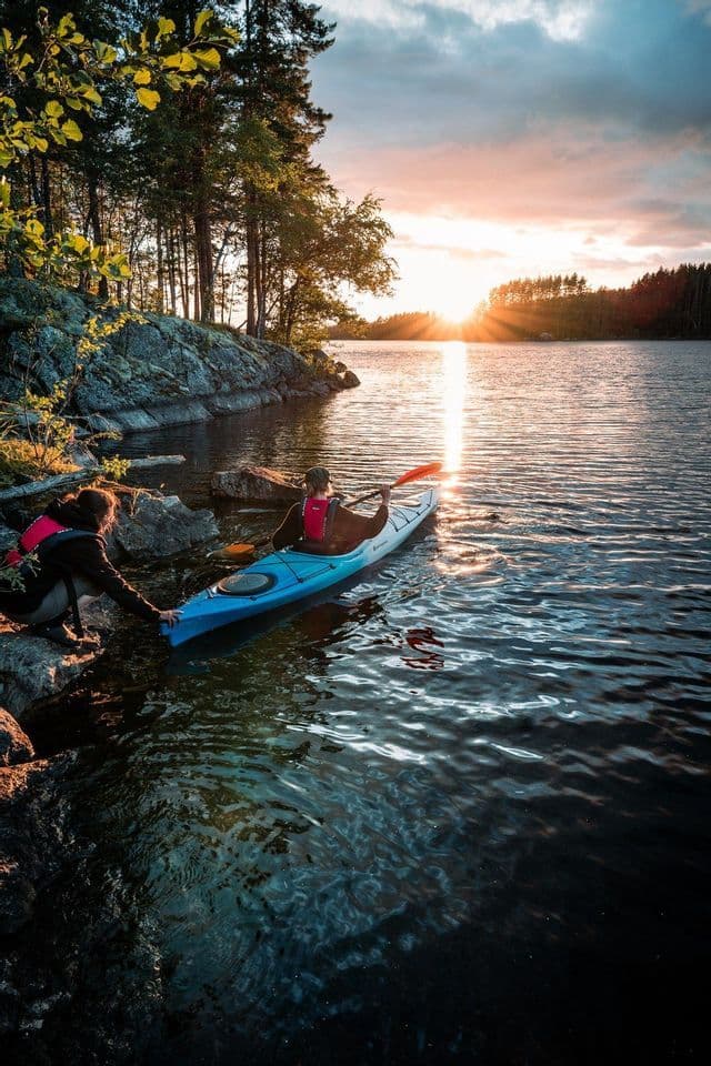 Eine Person einer WeRoad-Gruppenreise hilft einer anderen, ein Kajak von einem felsigen Ufer in einen ruhigen See bei Sonnenuntergang zu Wasser zu lassen.