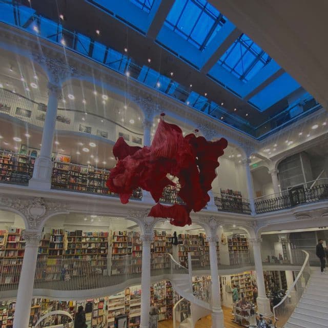 Una gran instalación de arte rojo cuelga del techo de una librería de varios niveles con columnas blancas ornamentadas y un tragaluz de cristal.