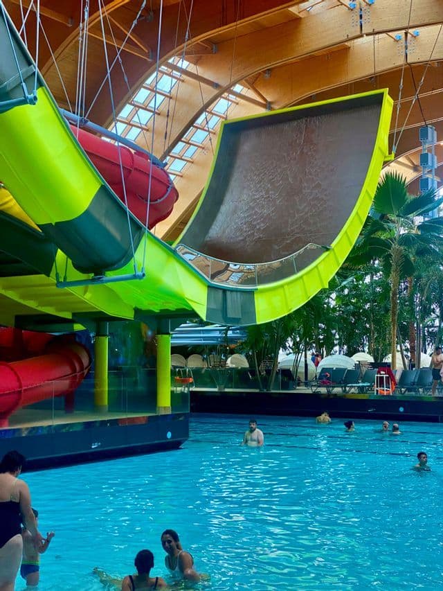 Grandes toboganes acuáticos verdes y rojos se elevan sobre una piscina llena de gente en un parque acuático cubierto.