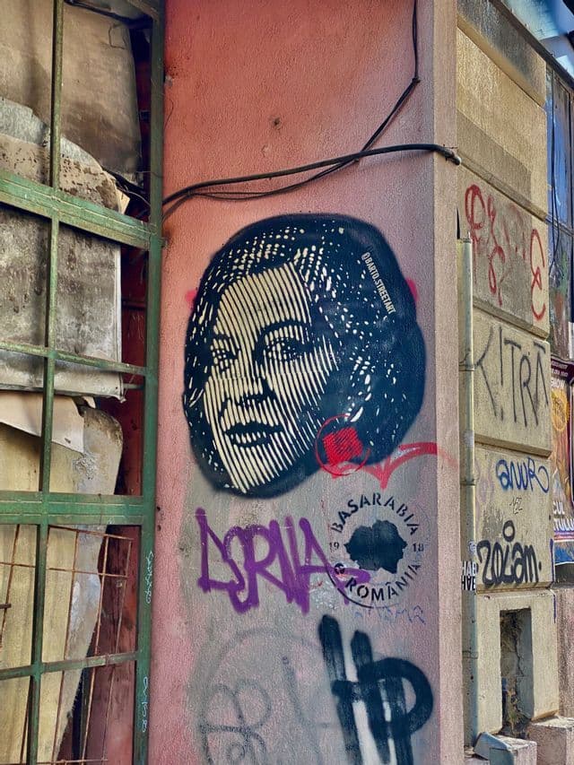 Un esténcil del rostro de una mujer pintado con aerosol en una pared urbana rosa, cubierta con varios grafitis coloridos.