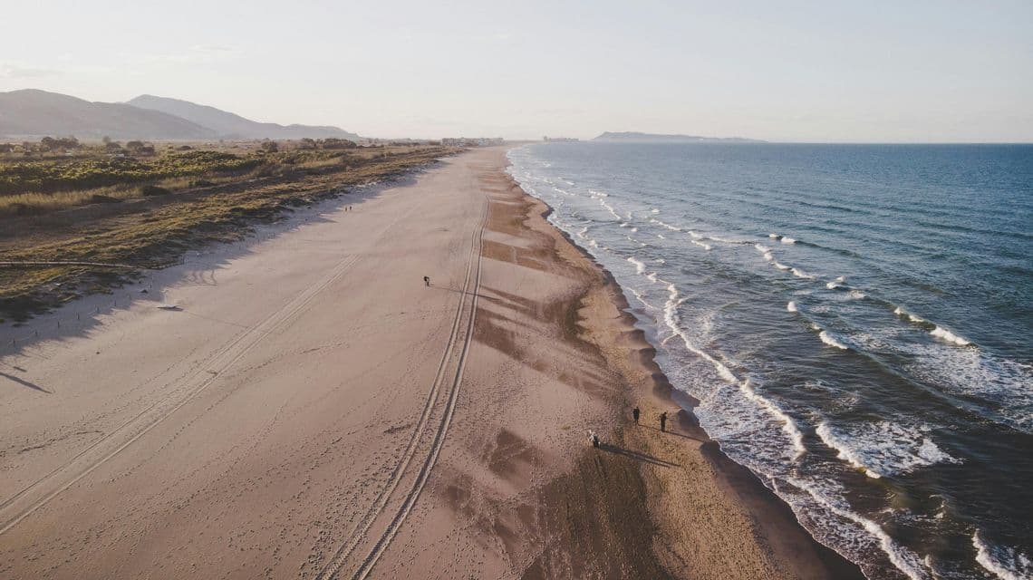 Veduta aerea di un gruppo WeRoad che cammina su un'ampia spiaggia sabbiosa lungo l'oceano, con montagne visibili in lontananza.