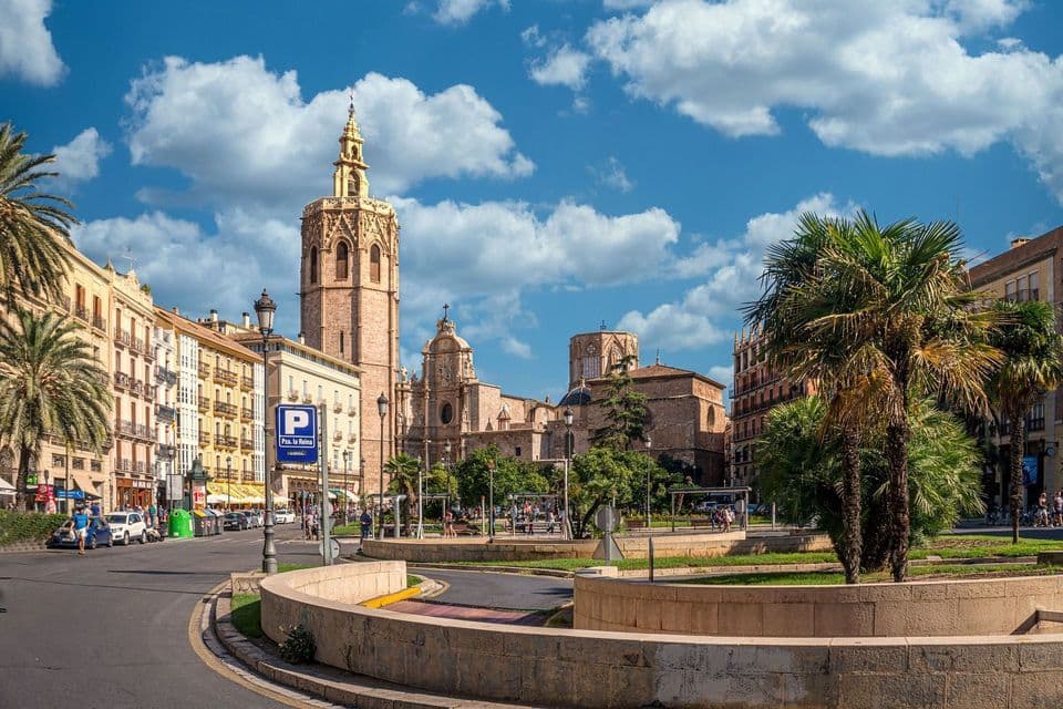 Una catedral de piedra con un campanario alto y ornamentado domina una soleada plaza de la ciudad con una rotonda y palmeras.