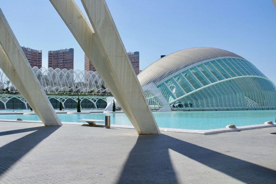 Un edificio moderno de cristal, con forma de cúpula, se encuentra junto a una piscina turquesa, con vigas arquitectónicas blancas en primer plano.