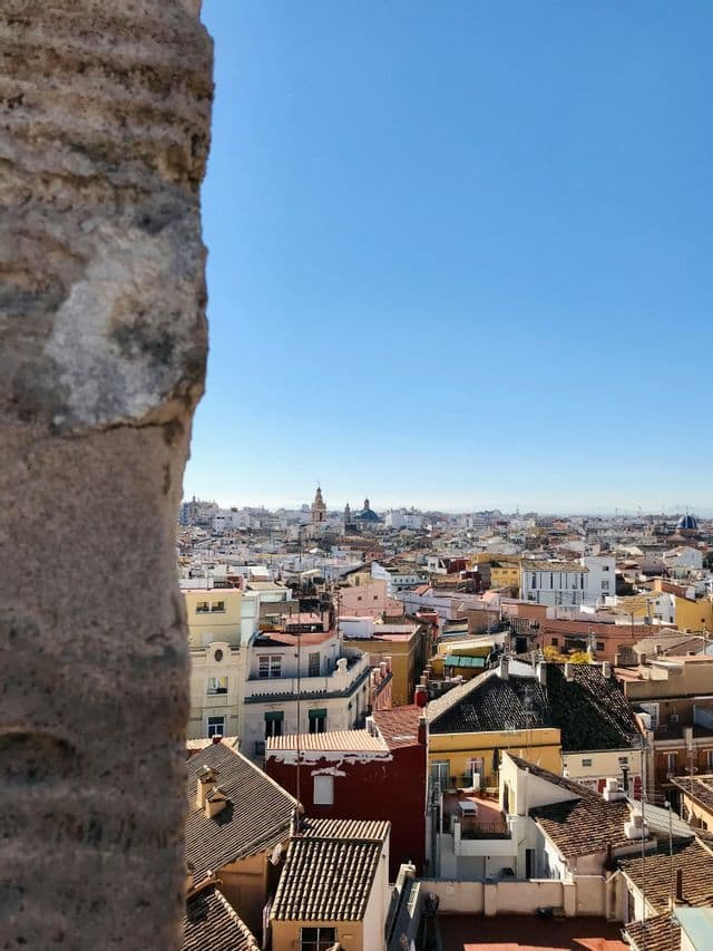 Una vista dall'alto di una città con edifici colorati e tetti in terracotta, incorniciata da un muro in pietra sotto un cielo azzurro limpido.