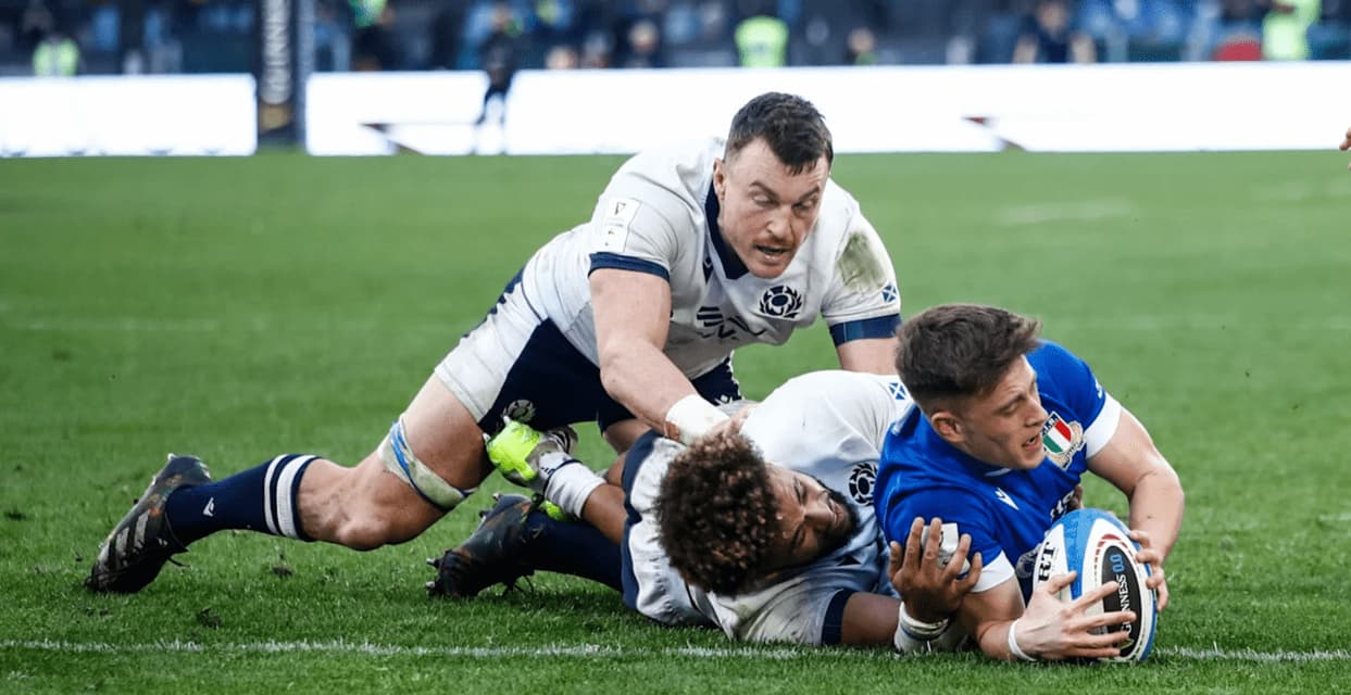 Un giocatore di rugby con una maglia blu segna una meta mentre viene placcato da due giocatori avversari su un campo d'erba.
