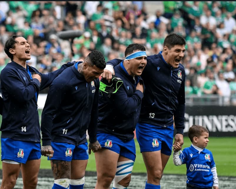 Quattro giocatori della nazionale italiana di rugby cantano sul campo, uno tiene per mano un bambino con la stessa maglia.