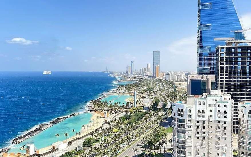 Una vista aerea di una moderna città costiera con grattacieli, spiagge orlate di palme e lagune turchesi accanto al mare blu profondo.