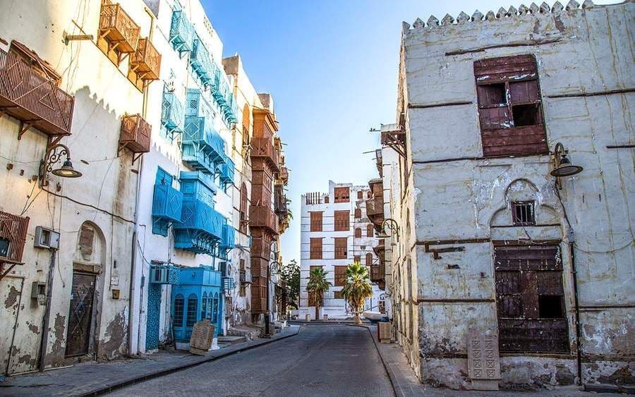 Una strada stretta e vuota è affiancata da tradizionali edifici bianchi con balconi in legno blu e marroni riccamente decorati sotto un cielo sereno.