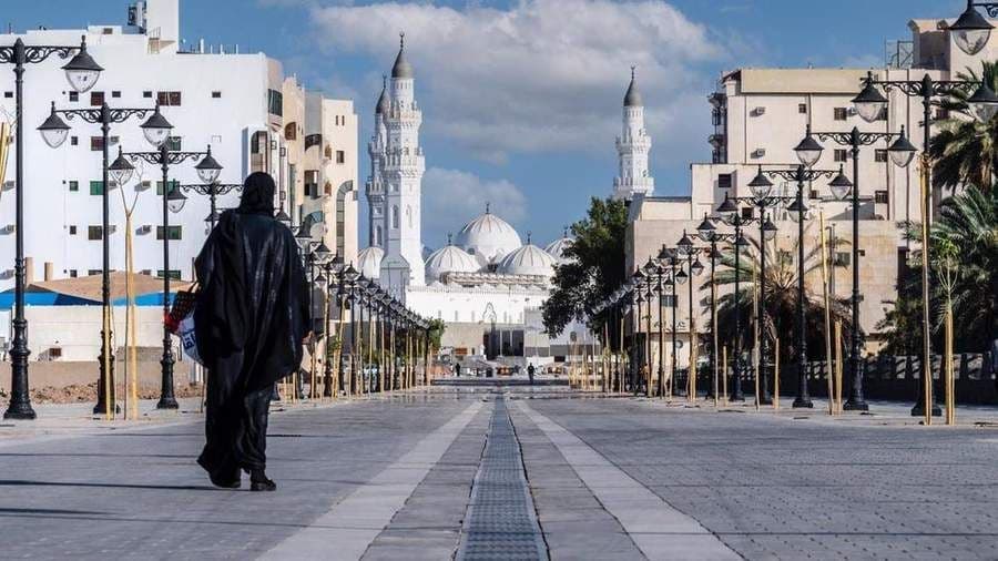 Una persona in abaya nero cammina lungo un'ampia strada fiancheggiata da lampioni verso una grande moschea bianca con minareti.