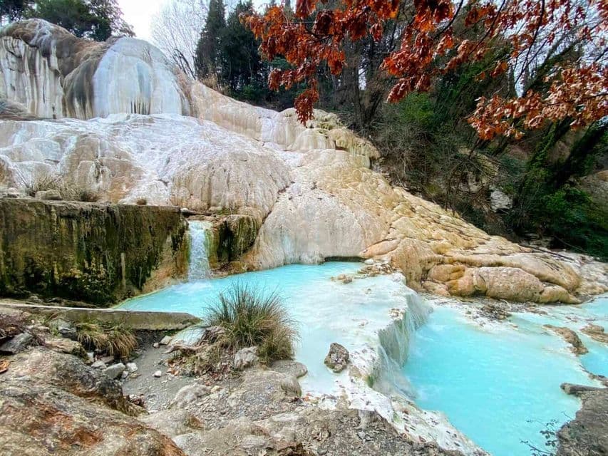 Terrazze di travertino bianco con piccole cascate che sfociano in piscine di acqua termale turchese, incorniciate da alberi con foglie autunnali.
