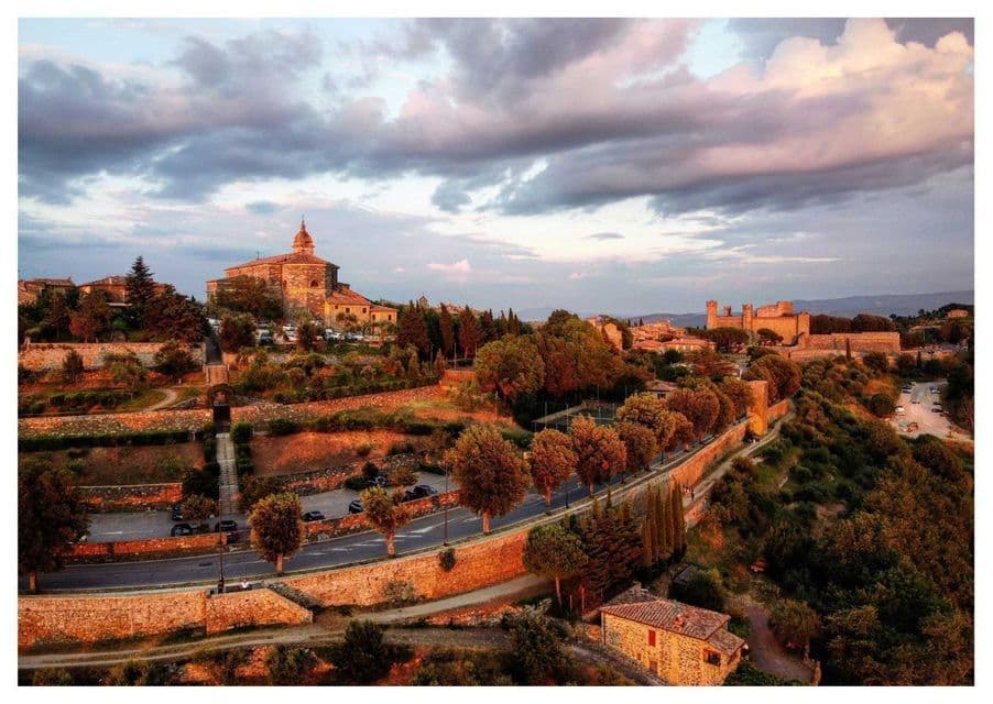 Una vista elevata di una città storica su una collina con edifici in pietra, una chiesa e una fortezza sotto un cielo nuvoloso al tramonto.