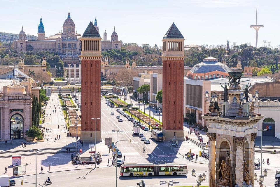 A sunny cityscape view of a wide avenue flanked by two large brick towers leading towards a palace on a hill.