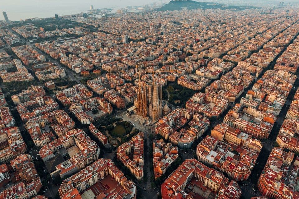An aerial view of the Sagrada Família cathedral surrounded by the grid-like streets and buildings of a sprawling city at sunset.