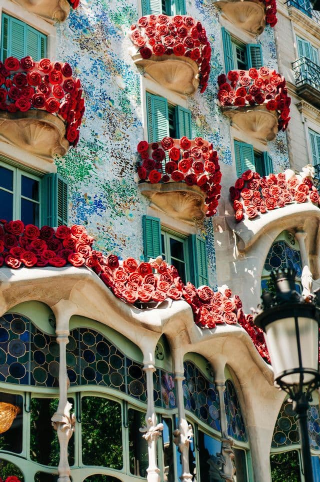An ornate building facade with colorful mosaic tiles, teal shutters, and balconies overflowing with bright red roses.