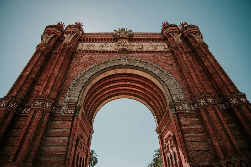 A low-angle shot of an ornate triumphal arch made of red brick with intricate sculptures against a clear blue sky.