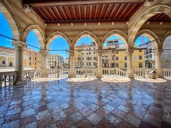 Una vista da una loggia in pietra con archi a sesto acuto e un pavimento a scacchi di marmo che si affaccia su una piazza cittadina soleggiata.