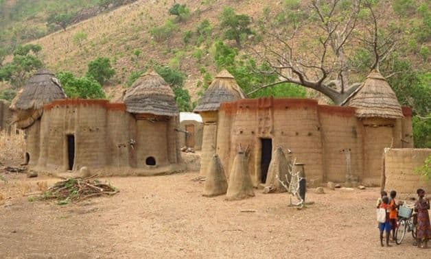 Capanne tradizionali in fango con tetti di paglia in un villaggio ai piedi di una collina verde, con alcuni bambini sulla destra.