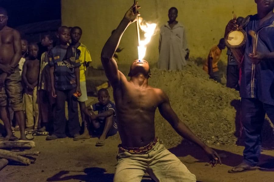 Un uomo a torso nudo si inginocchia a terra di notte, sputando fuoco da un bastone mentre una folla di persone osserva.