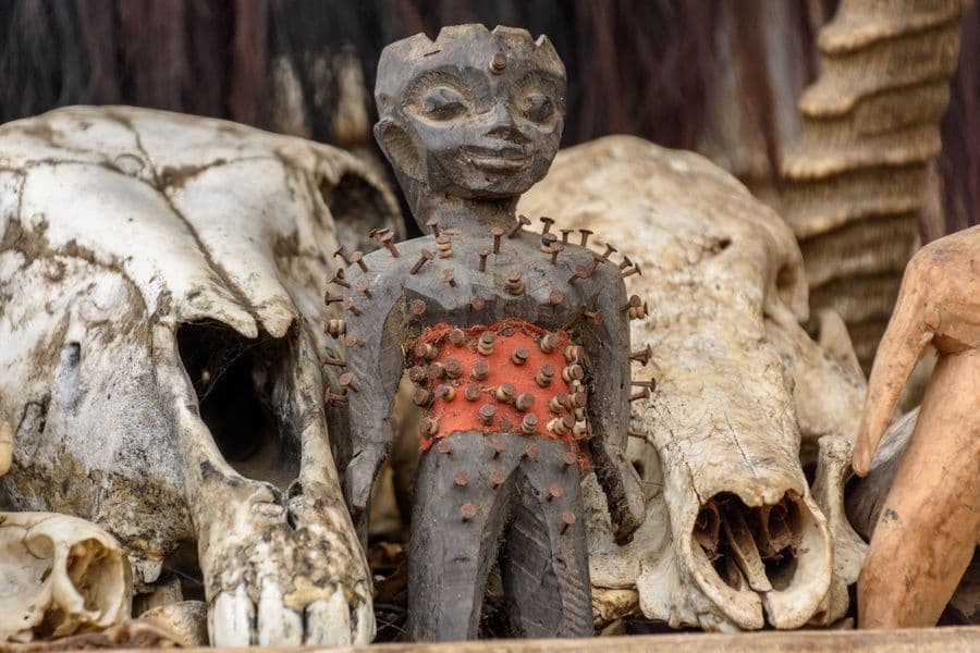 Un primo piano di una statuetta in legno intagliato, incastonata con chiodi arrugginiti, posizionata tra due grandi teschi di animali.