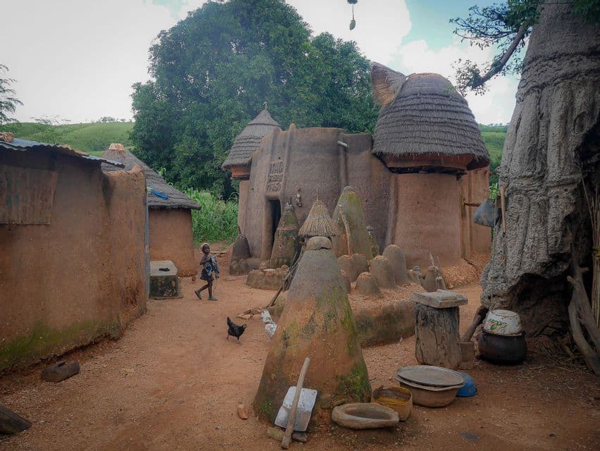 Un bambino cammina accanto a un pollo in un villaggio con capanne tradizionali di fango e tetti di paglia.