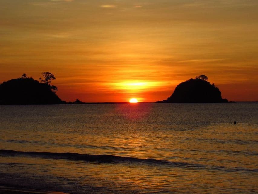 Il sole tramonta all'orizzonte sul mare, la sua luce arancione si riflette sull'acqua tra due isole in silhouette.