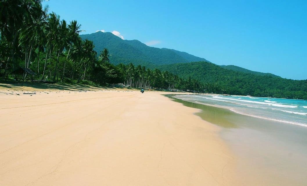 Una vasta spiaggia sabbiosa con onde turchesi che lambiscono la riva, costeggiata da palme ai piedi di una montagna verde.