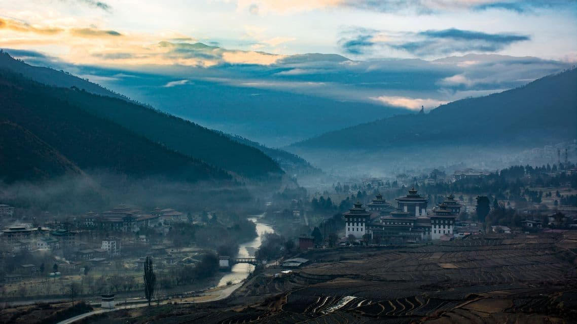 Ein Ort mit traditioneller Architektur und terrassierten Feldern liegt in einem nebligen Bergtal an einem Fluss bei Sonnenaufgang.