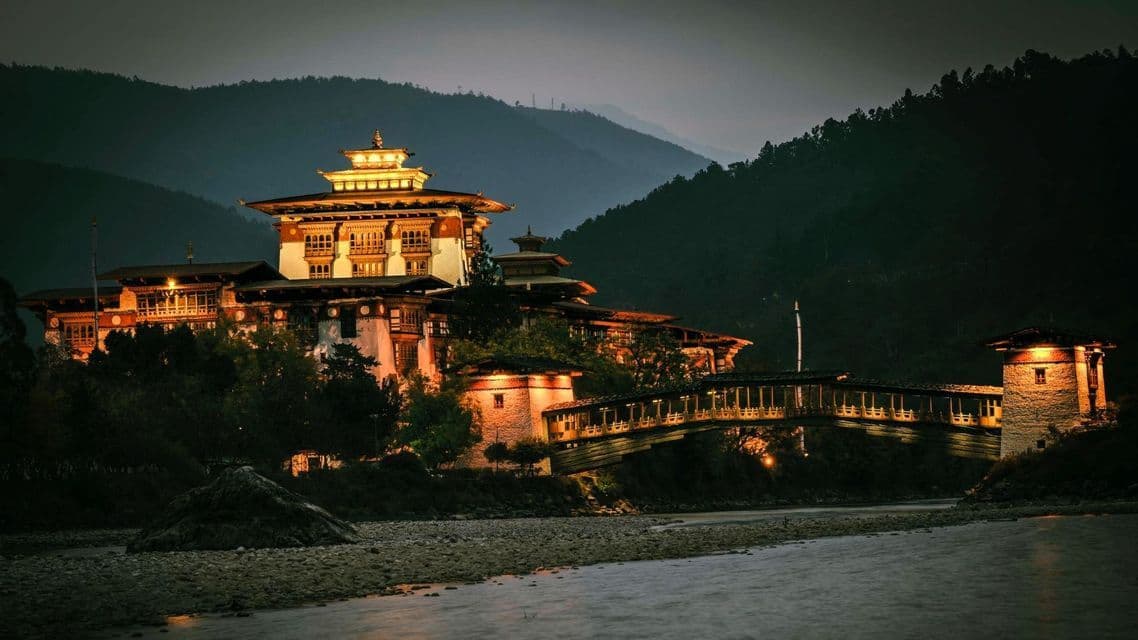 Ein beleuchtetes traditionelles Gebäude und eine überdachte Brücke an einem Fluss in der Abenddämmerung, mit Bergen im Hintergrund.