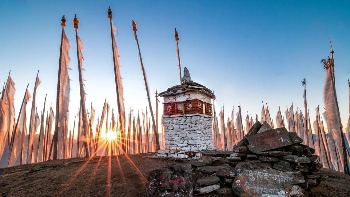Ein kleiner steinerner Stupa auf einem felsigen Hügel, umgeben von zahlreichen Gebetsfahnen, im goldenen Licht des Sonnenaufgangs.