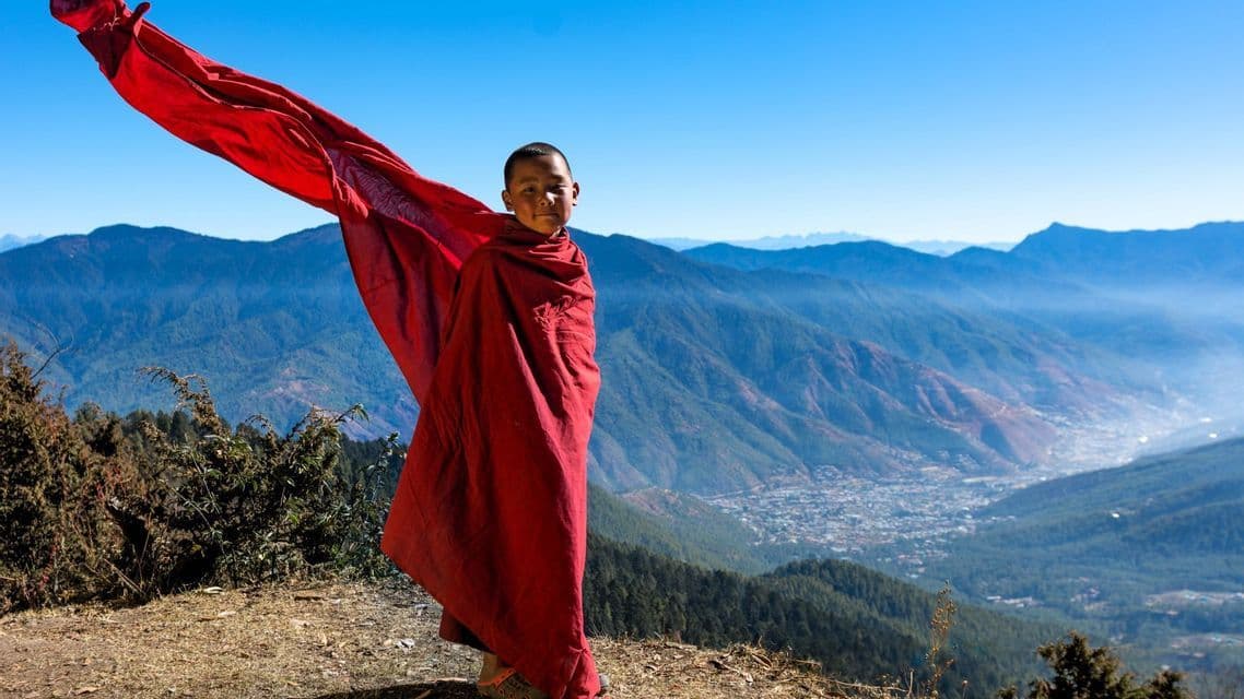 Ein junger Junge in roten Gewändern steht auf einem Berggipfel, sein Gewand weht im Wind über einem Tal.
