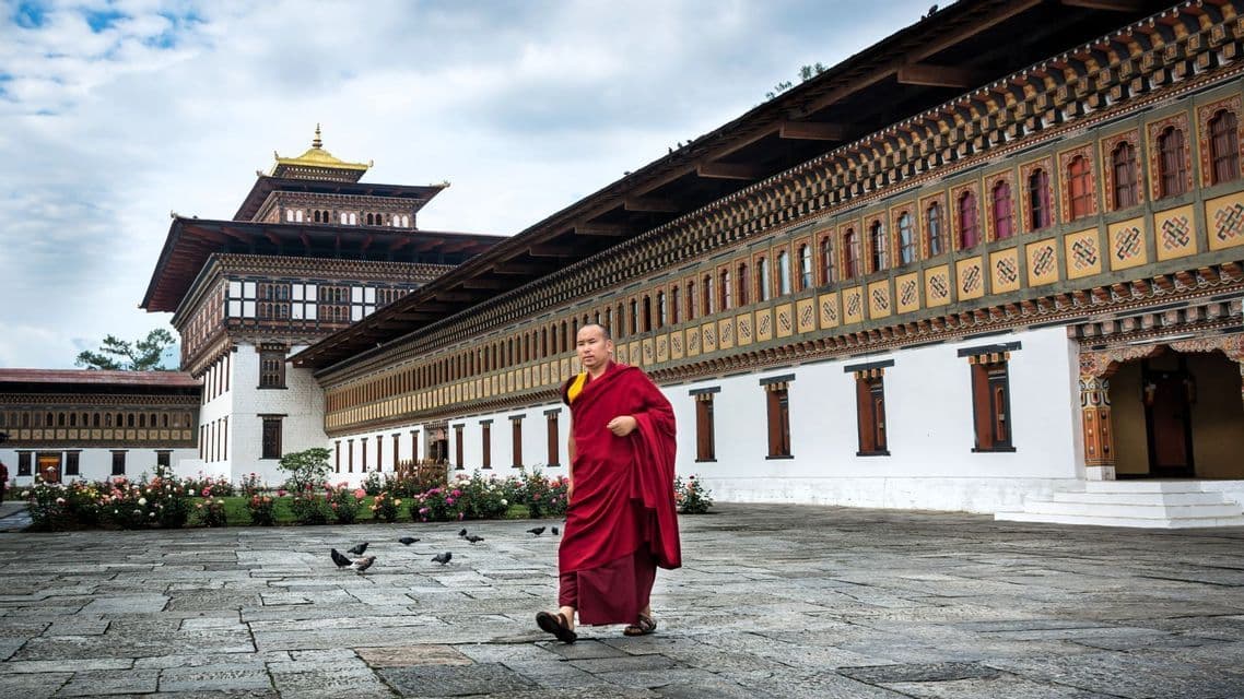 Ein buddhistischer Mönch in roten Roben geht über einen steinernen Hof vor einem verzierten Kloster mit goldenem Dach.
