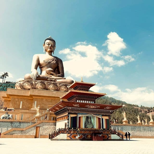Eine massive bronzene Buddha-Statue sitzt auf einem Thron und blickt auf einen kleineren, verzierten Tempel in einem großen Innenhof.