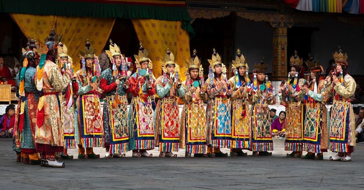 Eine Reihe von Darstellern in farbenfrohen traditionellen Gewändern und aufwendigen goldenen Kopfbedeckungen während eines kulturellen Festivals im Freien.