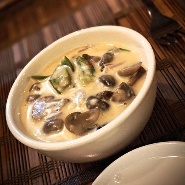 Eine weiße Schale cremiger Pilzsuppe mit grünem Gemüse, serviert auf einem geflochtenen Bambus-Tischset.