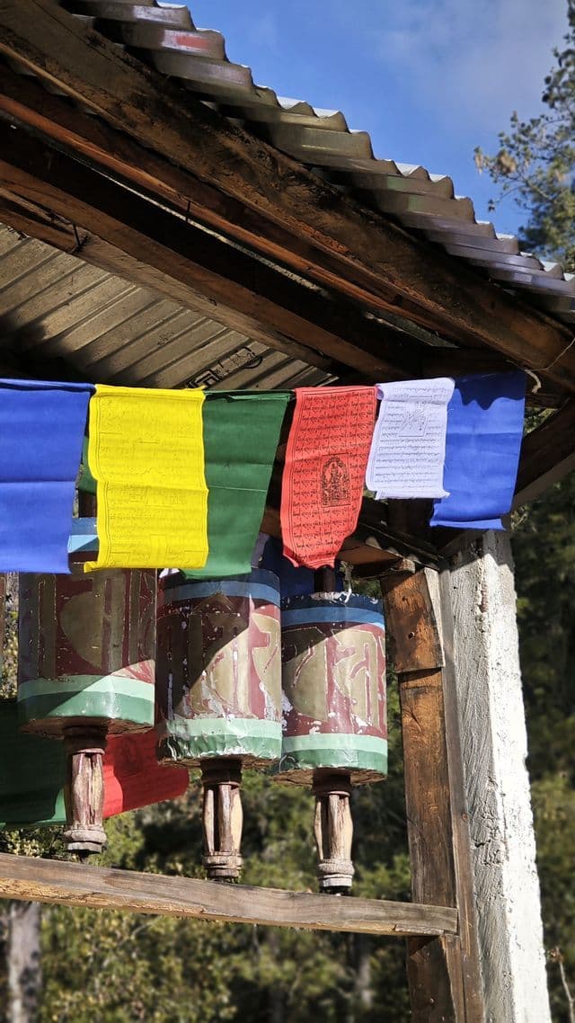 Eine Reihe tibetischer Gebetsmühlen mit bunten Gebetsfahnen, die darüber unter einem rustikalen Holz- und Wellblechdach hängen.