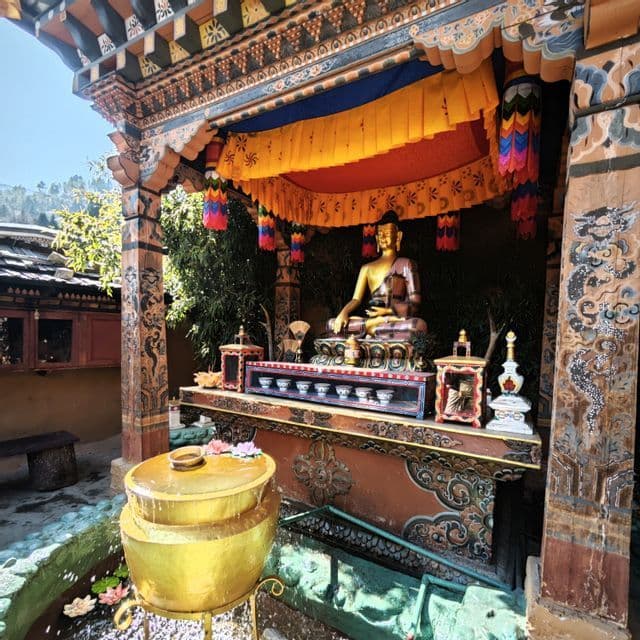 Eine goldene Buddha-Statue thront auf einem geschmückten Altar in einem verzierten Holzschrein mit einem gelb-roten Baldachin und einem Brunnen im Vordergrund.