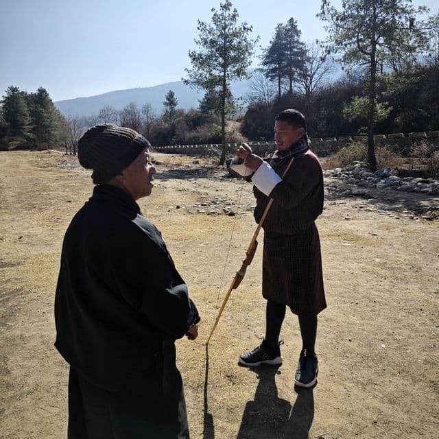 Ein Mann in traditioneller Tracht, der einen Holzbogen hält, spricht mit einer anderen Person, die eine Mütze trägt, auf einem sonnigen Feld mit Bäumen im Hintergrund.