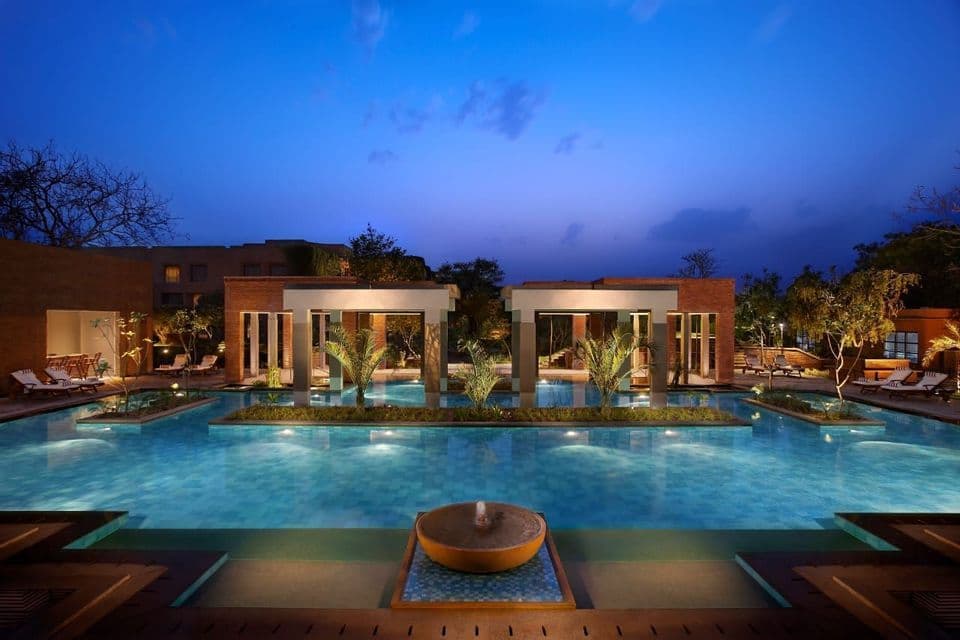 Ein Resort-Swimmingpool mit Unterwasserbeleuchtung, die in der Dämmerung zwei zentrale Pavillons und einen Springbrunnen beleuchtet.