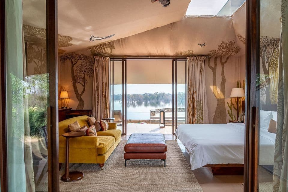 Una lujosa habitación tipo tienda de campaña con un sofá amarillo y una cama, con puertas de cristal que se abren a una serena vista al lago.