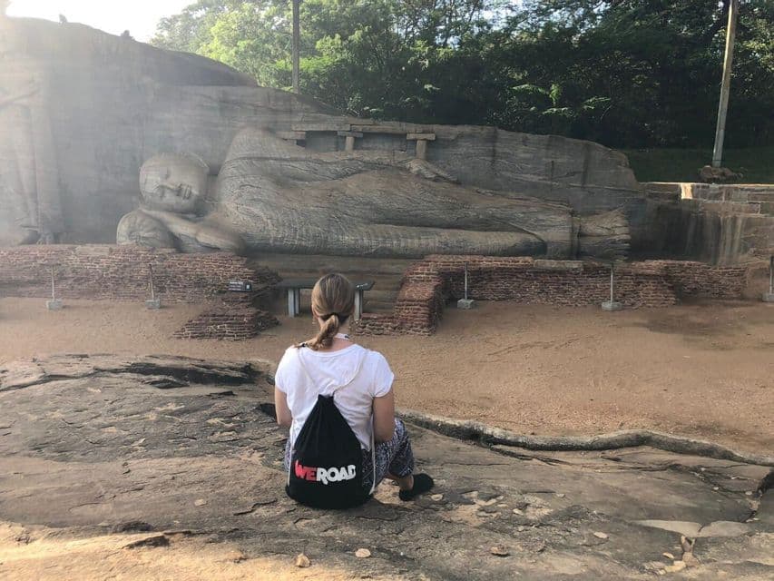 Une femme avec un sac à dos WeRoad est assise sur un rocher, observant une immense statue de Bouddha couché sculptée dans une falaise.