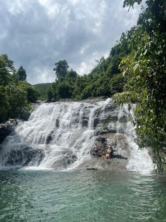 Un gruppo WeRoad in posa in costume sulle rocce di un'ampia cascata, circondata da una rigogliosa foresta verde.