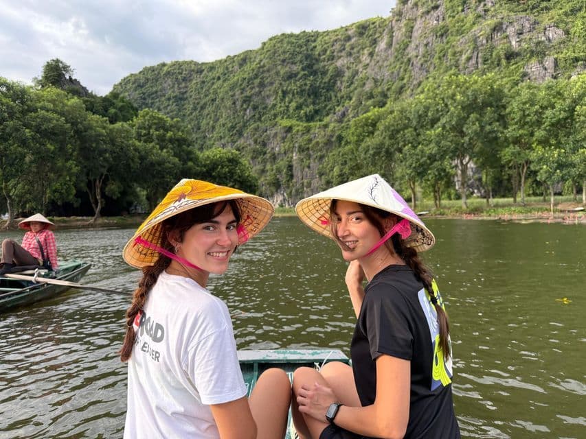 Due viaggiatrici di un gruppo WeRoad, con cappelli conici, sorridenti su una barca circondata da rigogliose montagne verdi.