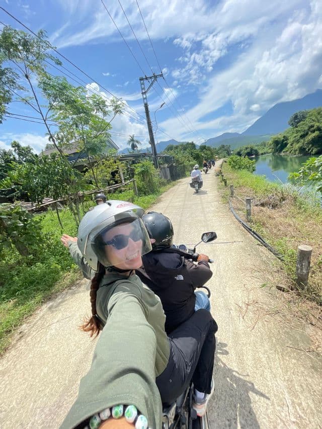 Una donna con il casco si scatta un selfie dal retro di una moto durante un viaggio di gruppo WeRoad su una strada rurale accanto a un fiume.