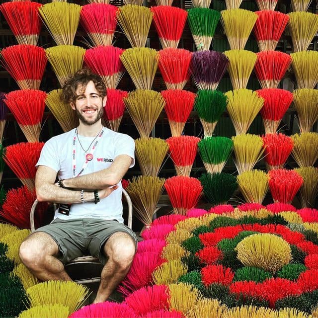 Un uomo sorridente con un cordino WeRoad è seduto su una sedia davanti a un muro di fasci di bastoncini di incenso colorati.