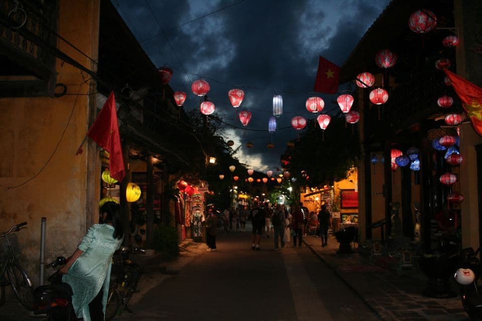 Una strada stretta di notte è illuminata da lanterne di carta rosse e blu appese tra gli edifici mentre le persone camminano sotto.