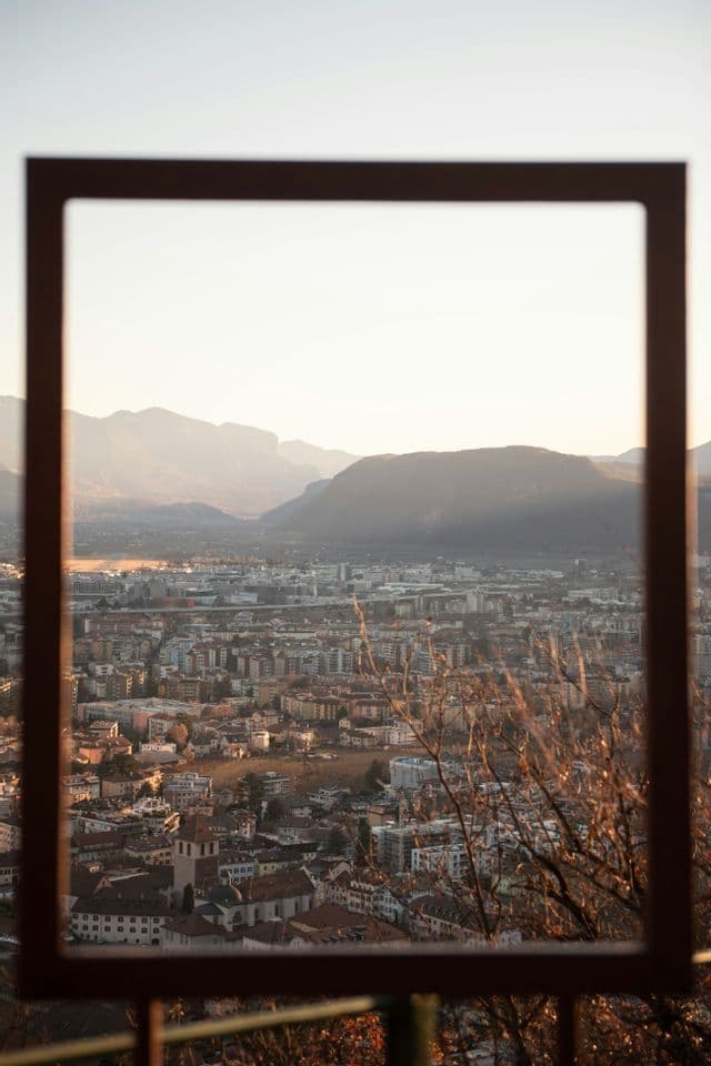 Un paesaggio urbano in una valle di montagna, visto attraverso una grande cornice rettangolare in primo piano, illuminato da calda luce solare.