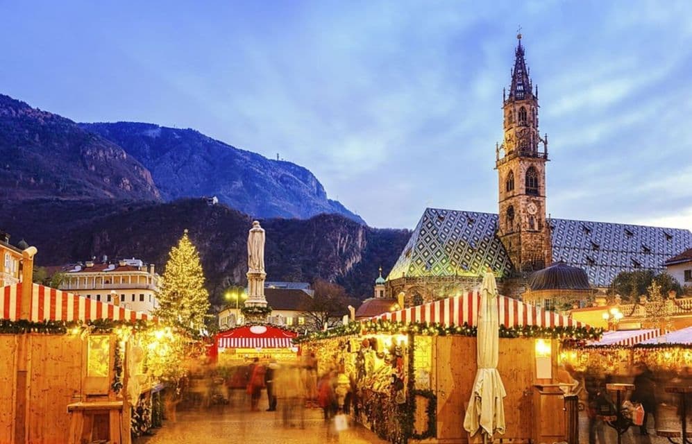Un vivace mercatino di Natale al tramonto con bancarelle illuminate in una piazza cittadina, con una grande cattedrale e montagne sullo sfondo.