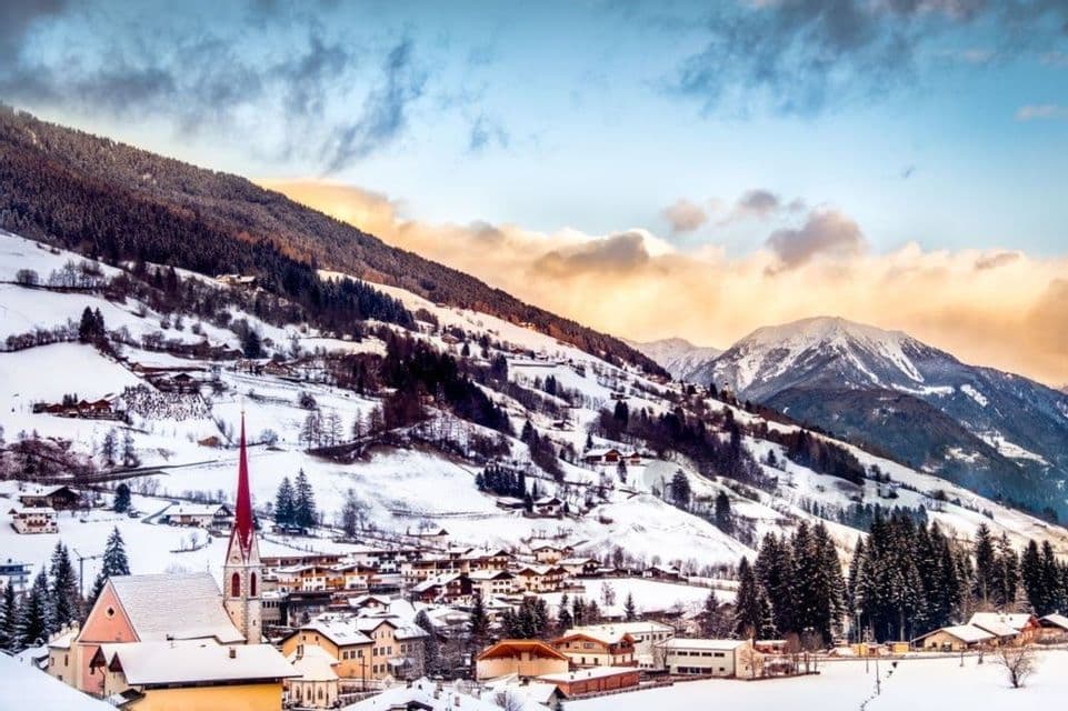 Un villaggio con una chiesa dal campanile rosso prominente è adagiato in una valle di montagna innevata, sotto un cielo colorato.