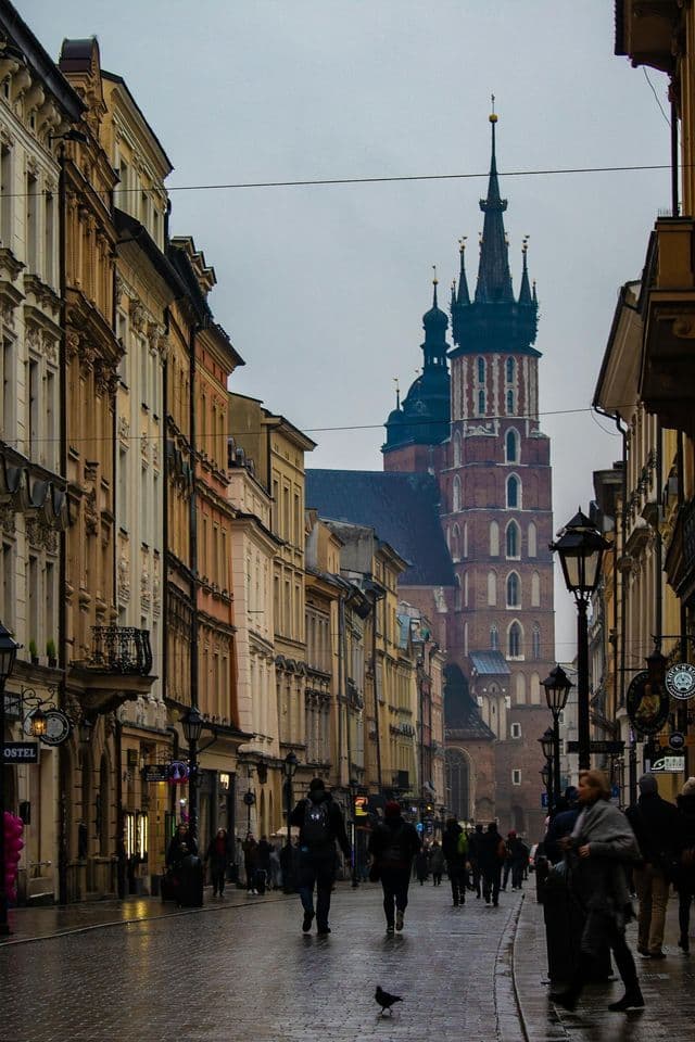 Persone camminano lungo una strada acciottolata bagnata, fiancheggiata da edifici storici, dirette verso una grande chiesa di mattoni rossi con guglie, sotto un cielo coperto.